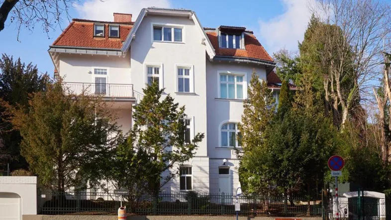 Klassische Villa mit weißer Fassade, rotem Dach und umgebenden Bäumen in sonniger Lage.