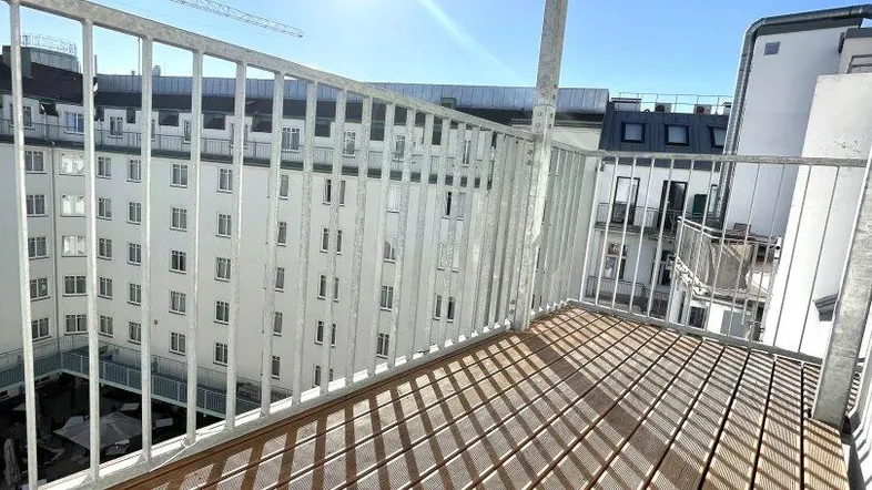 Sonniger Balkon mit Holzboden und Blick auf umliegende Gebäude unter blauem Himmel.