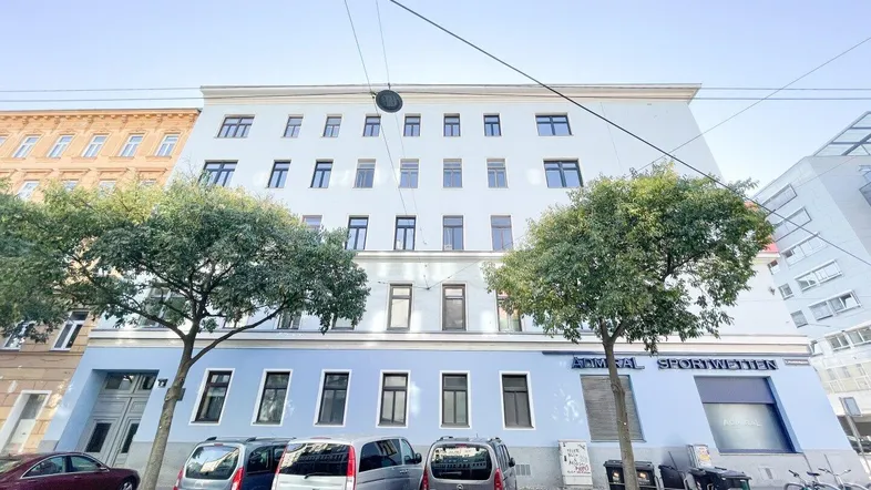 Gebäudefassade mit hellblauer und weißer Farbgebung, umgeben von Bäumen und parkenden Autos auf der Straße.