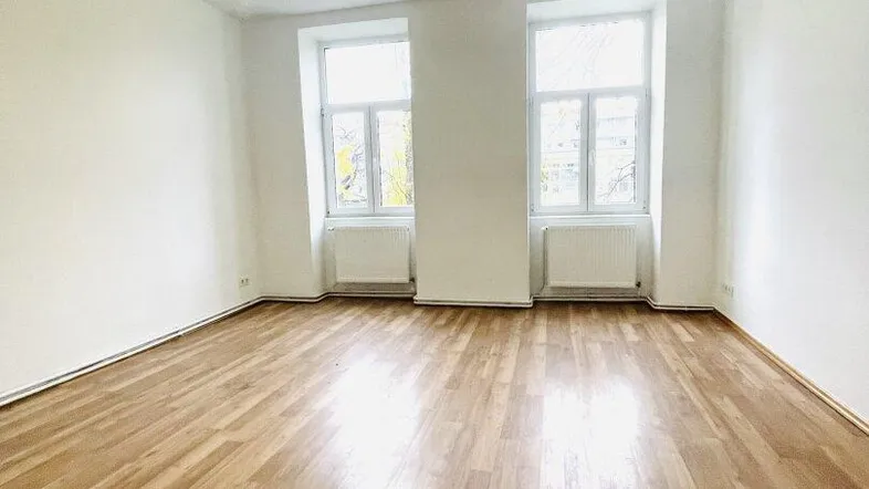 Helles, leeres Zimmer mit Parkettboden und zwei großen Fenstern, bereit zur individuellen Gestaltung.
