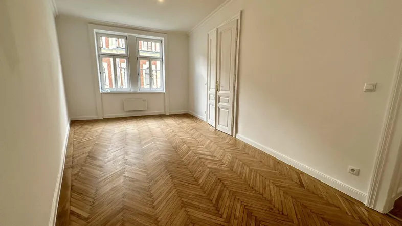 Helles Zimmer mit Fischgrätparkett und hohen Fenstern, ideal als Wohn- oder Schlafzimmer.