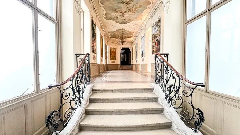 Prunkvoller Eingangsbereich mit kunstvollen Deckenmalereien, geschwungener Treppe und schmiedeeisernem Geländer.
