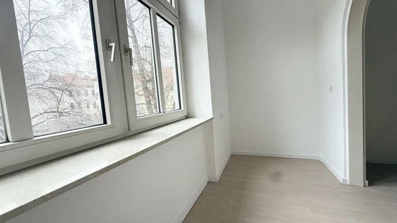 Helles, leeres Zimmer mit großen Fenstern, hellem Boden und einem eleganten Torbogen.