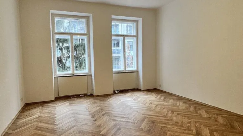 Heller Raum mit klassischem Fischgrätparkett und zwei großen Fenstern, ideal als Wohnzimmer.