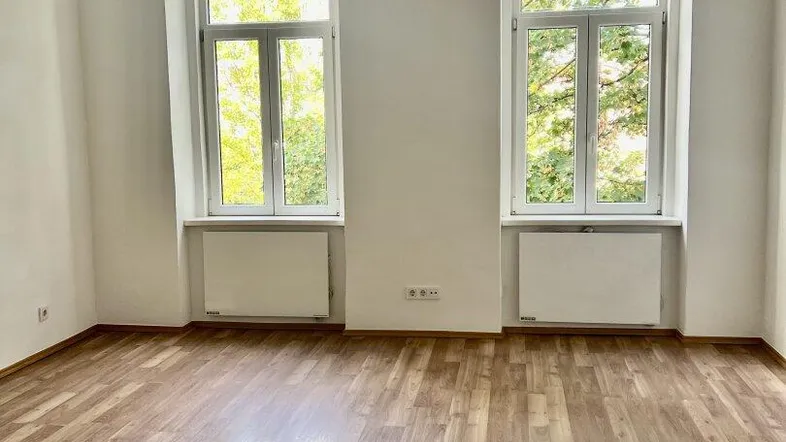 Heller Wohnraum mit zwei großen Fenstern, Laminatboden und Blick ins Grüne.