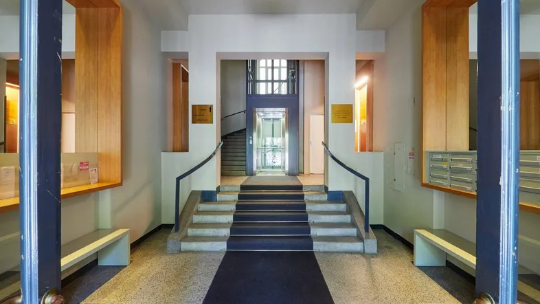 Heller Eingangsbereich mit Terrazzoboden, Treppe und modernem Glasaufzug im Hintergrund.