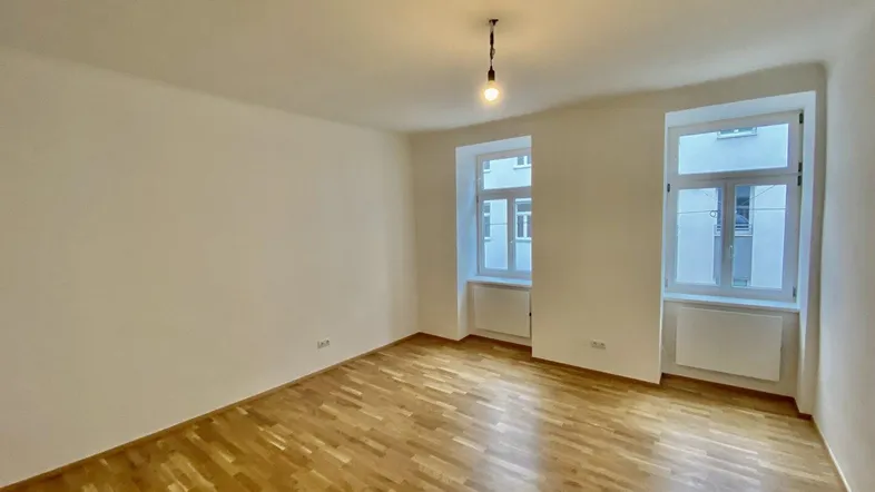 Helles, leeres Zimmer mit Parkettboden und zwei Fenstern, bereit zur individuellen Gestaltung.