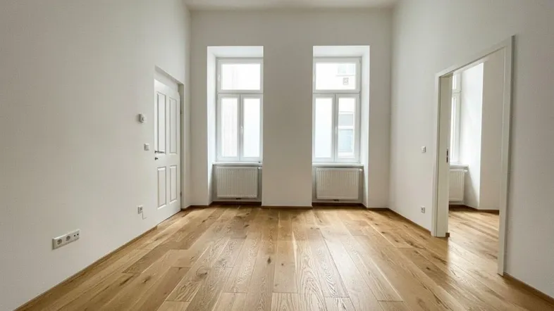 Helles Zimmer mit neuem Holzfußboden, weißen Wänden und hohen Fenstern, bereit zur individuellen Gestaltung.
