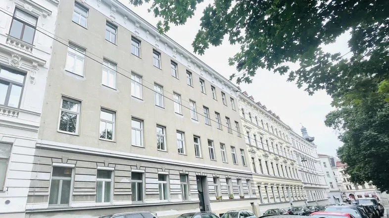Klassisches Mehrfamilienhaus mit heller Fassade und parkenden Autos auf der Straße.