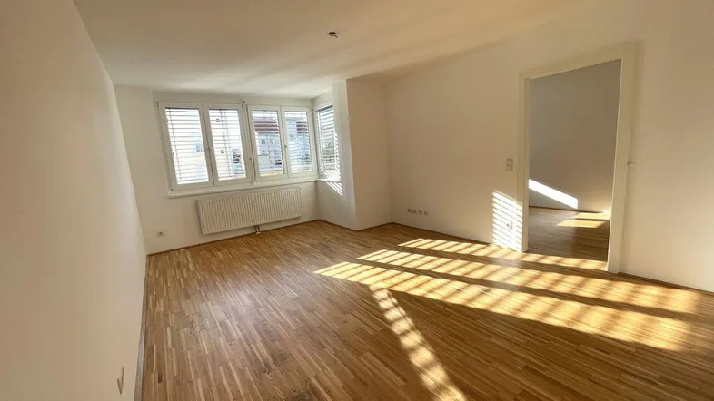 Helles Zimmer mit Parkettboden, weißen Wänden und Fenster mit Jalousien, durchflutet von Sonnenlicht.