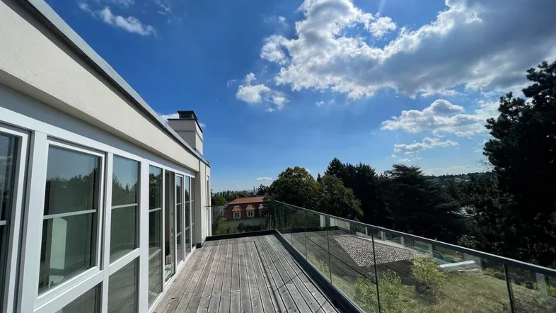 Sonnige Terrasse mit Holzboden, Glasgeländer und weitem Blick über die grüne Umgebung.