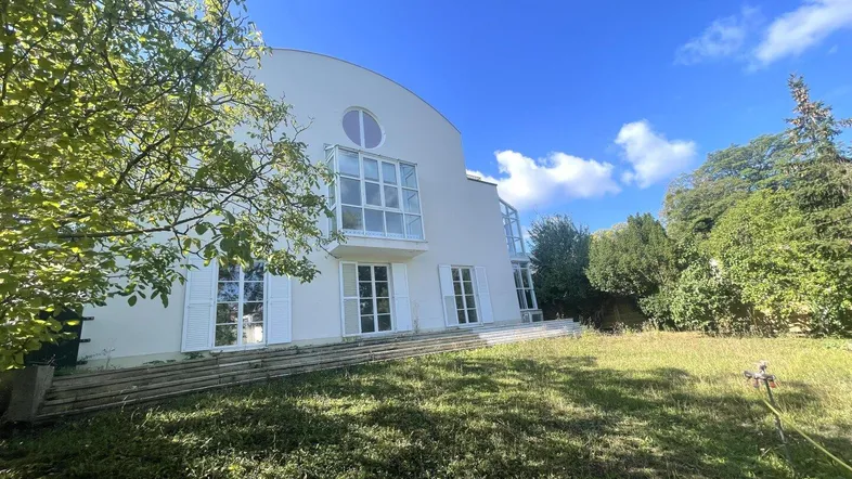 Modernes weißes Gebäude mit großen Fenstern und umliegendem grünen Garten unter blauem Himmel.