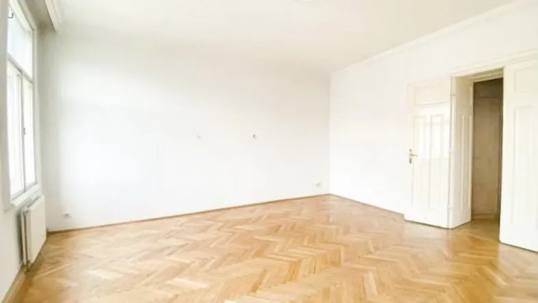 Helles, leeres Zimmer mit Fischgrätparkett, weißen Wänden und Stuckdecke.