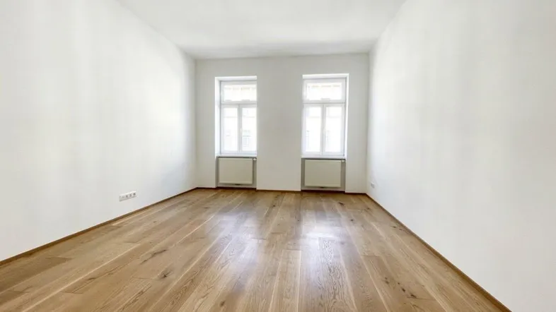 Helles, leeres Zimmer mit Holzboden und zwei Fenstern, bereit zur individuellen Gestaltung.