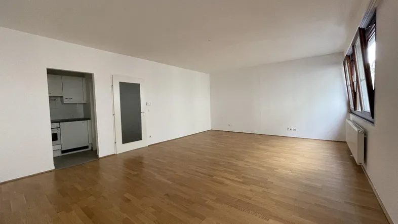 Geräumiges Wohnzimmer mit hellem Parkettboden und Zugang zu einer kleinen Küchenzeile.