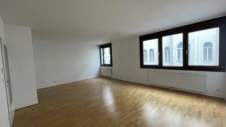Helles, leeres Wohnzimmer mit Parkettboden und großen Fenstern, die viel Tageslicht hereinlassen.