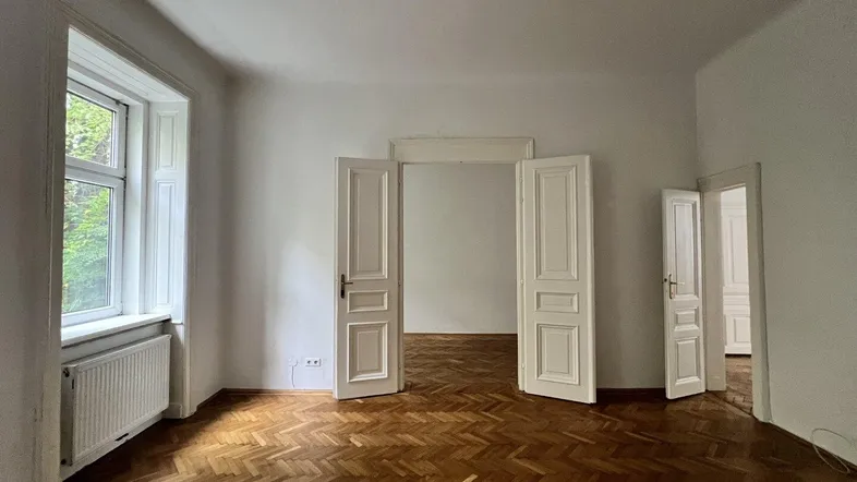 Helles, leeres Zimmer mit Fischgrätparkett und hohen Flügeltüren, bereit zur individuellen Gestaltung.