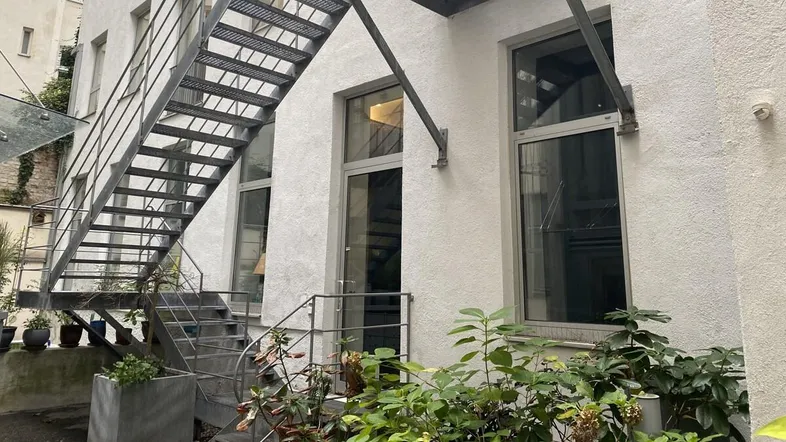 Außenansicht eines Gebäudes mit weißer Fassade, Metalltreppe und Pflanzen im begrünten Hofbereich.