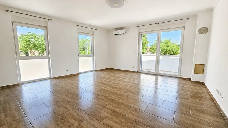 Helles Wohnzimmer mit Fliesenboden, großen Fenstern und Zugang zu einem Balkon.