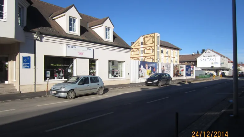 Straßenansicht mit Gebäuden, geparkten Autos und Geschäften unter blauem Himmel.