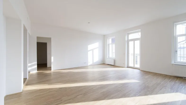 Helles, leeres Wohnzimmer mit Parkettboden, weißen Wänden und großen Fenstern mit viel Tageslicht.
