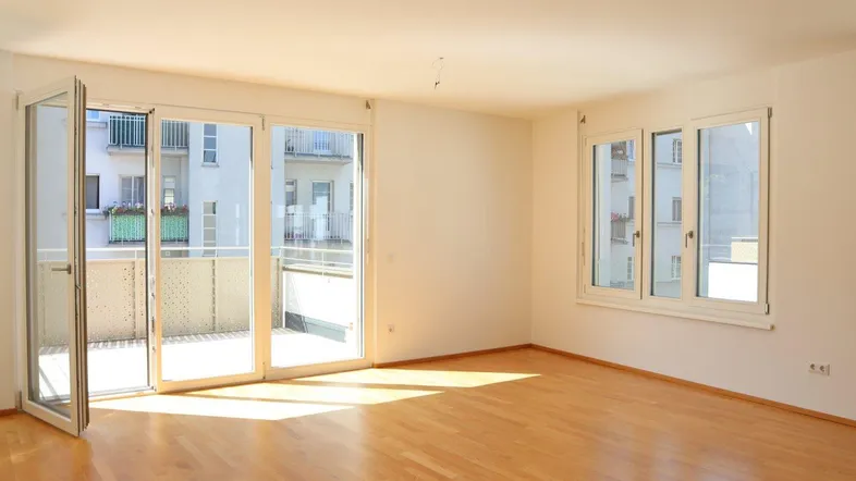 Helles Wohnzimmer mit Parkettboden, großen Fenstern und Zugang zu einem Balkon.