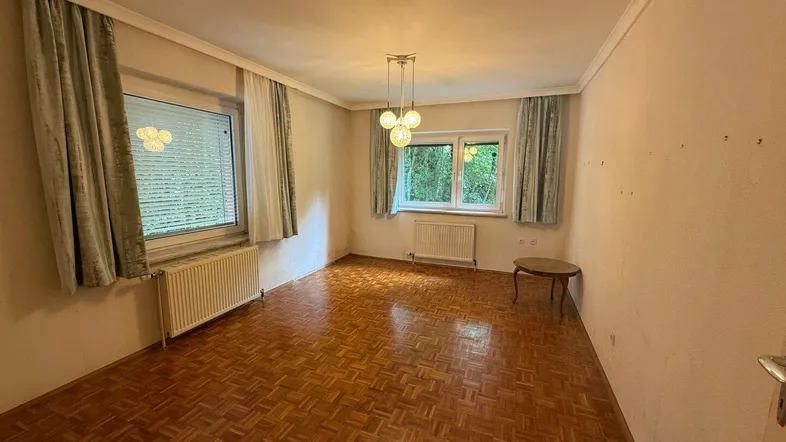 Helles Wohnzimmer mit Parkettboden, zwei Fenstern und schlichten Vorhängen, bietet eine grüne Aussicht.