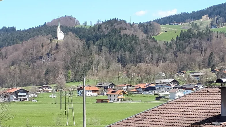 Panoramablick über eine ländliche Siedlung mit grünen Feldern, bewaldeten Hügeln und einer Kirche.