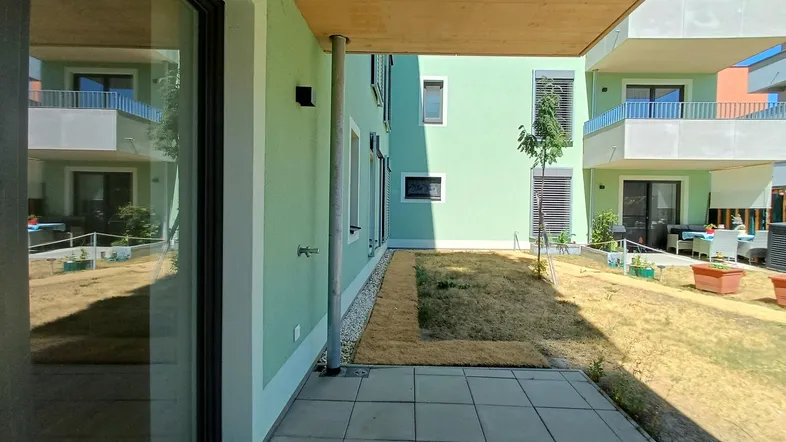 Eine überdachte Terrasse mit Zugang zu einem kleinen Gartenbereich in einer modernen Wohnanlage.