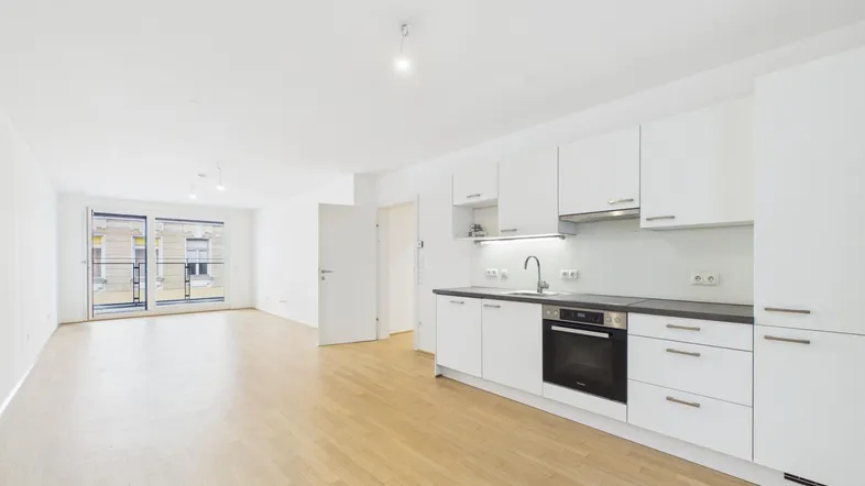 Heller offener Wohnbereich mit integrierter Küche, Parkettboden und Zugang zum Balkon.