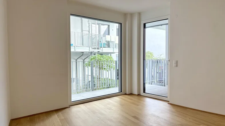 Helles Zimmer mit Parkettboden und großen Fenstern, die Zugang zu einem Balkon bieten.