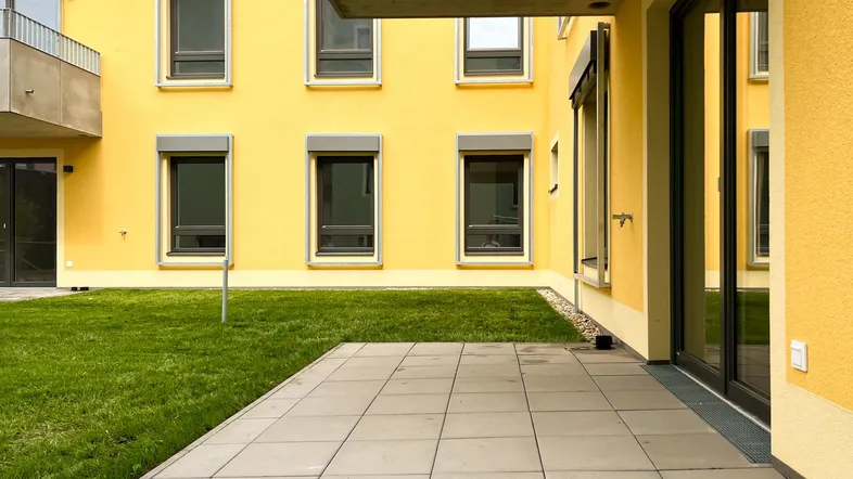 Eine gepflasterte Terrasse mit Rasenfläche vor einem modernen Gebäude mit gelber Fassade.