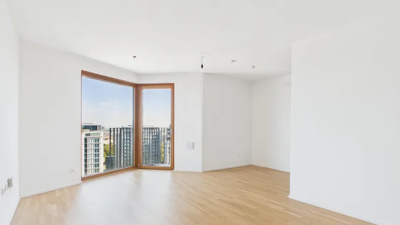 Helles, leeres Wohnzimmer mit Parkettboden und großen Fenstern, die einen Stadtblick bieten.