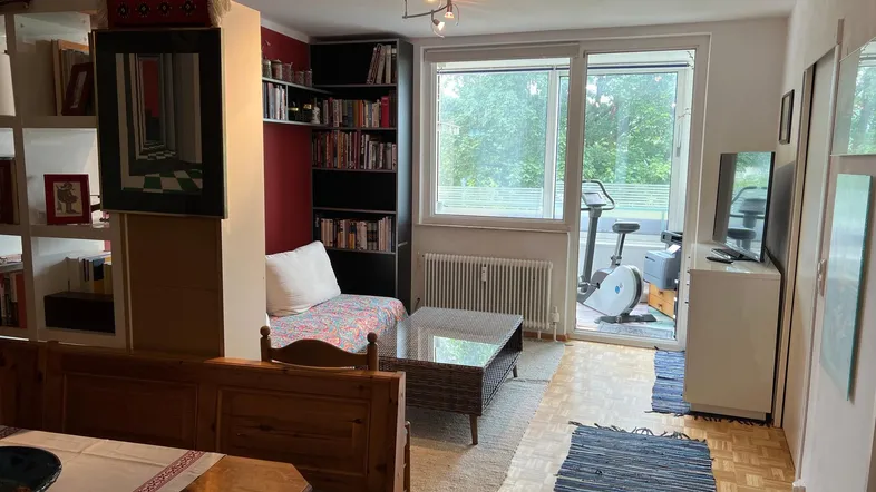 Gemütlicher Wohn- und Essbereich mit Holzbänken, Bücherregalen und Zugang zum Balkon mit Blick ins Grüne.