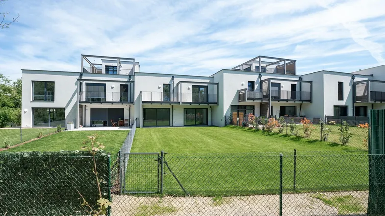 Moderne Reihenhäuser mit weißer Fassade, großen Fenstern, Balkonen und Dachterrassen, umgeben von einer grünen Rasenfläche.