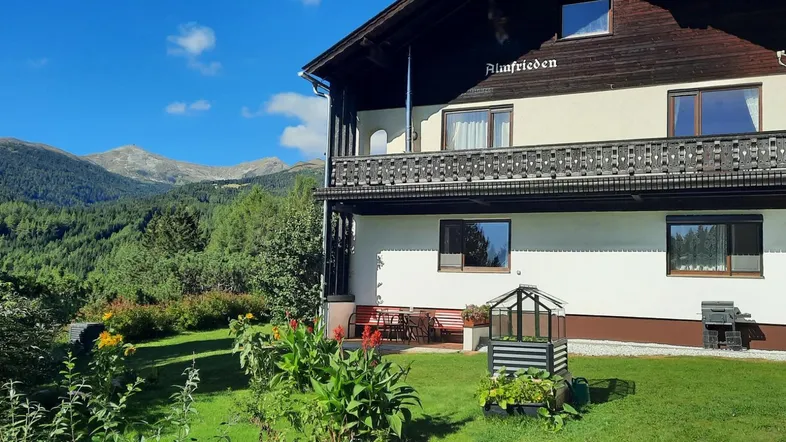 Haus mit Holzbalkon und Bergblick, umgeben von grüner Natur und klarem Himmel.