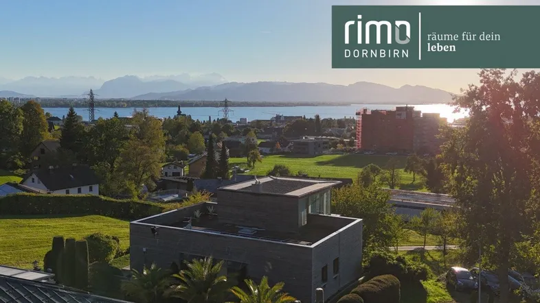 Panoramablick über eine Wohnsiedlung mit See und Bergen im Hintergrund bei Sonnenuntergang.