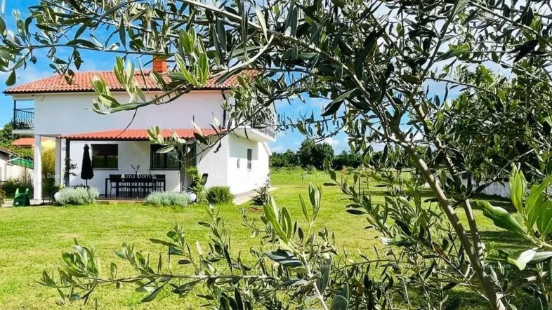 Weißes Haus mit rotem Ziegeldach, umgeben von einem grünen Garten mit Olivenbäumen und einer Terrasse.