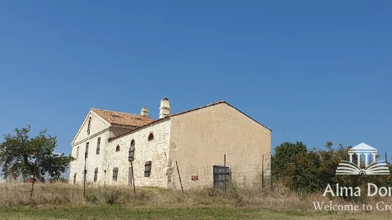 Altes Steinhaus mit Ziegeldach unter klarem blauem Himmel, umgeben von trockener Vegetation.