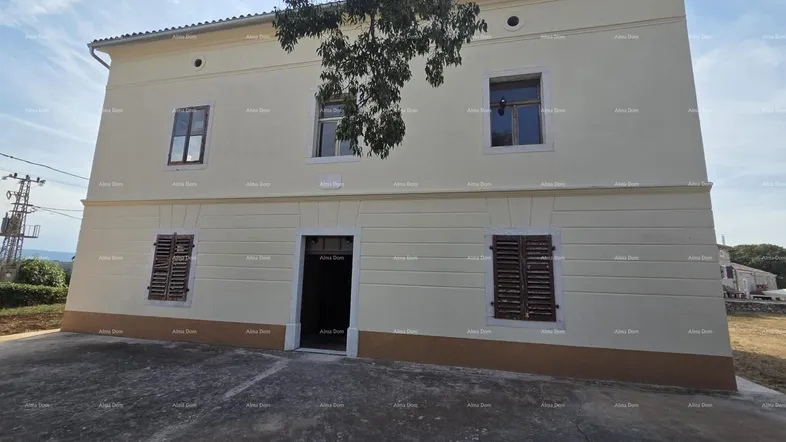 Fassade eines zweistöckigen Gebäudes mit hellbeigem Putz und braunem Sockel, umgeben von Natur.