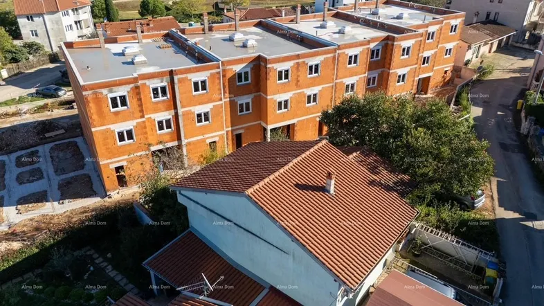 Luftaufnahme eines im Bau befindlichen Gebäudes mit orangefarbenen Ziegeln und einem älteren Haus im Vordergrund.