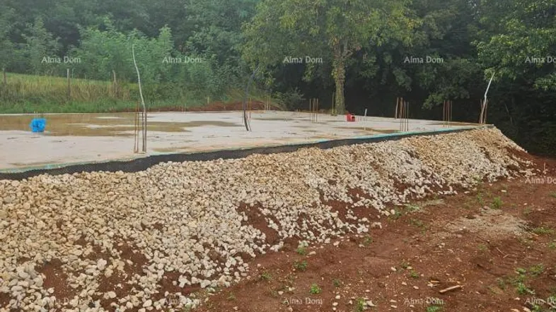 Betonfundament einer Baustelle mit Schotter und Erde, umgeben von Bäumen und ländlicher Umgebung.