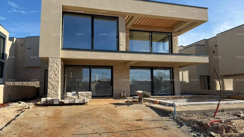 Modernes Reihenhaus im Bau mit großen Fenstern und beiger Fassade, umgeben von Baustellenmaterialien.