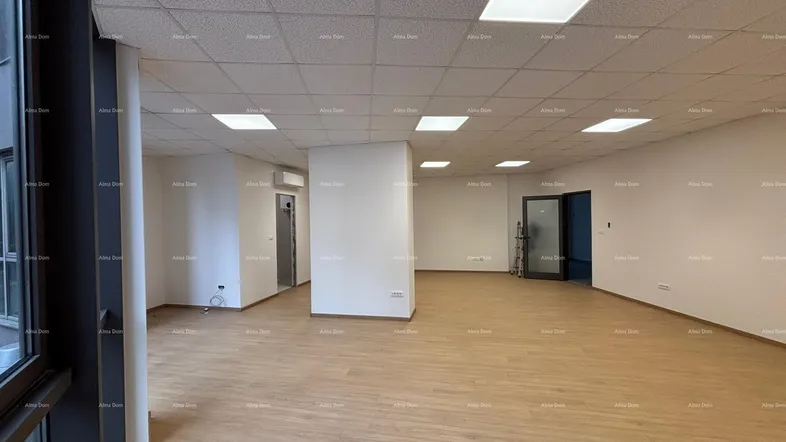 Großer, leerer Gewerberaum mit hellem Laminatboden, abgehängter Decke und großen Fenstern für viel Tageslicht.