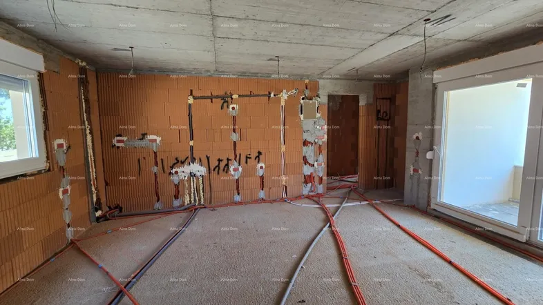 Großer Raum im Rohbau mit zwei Fenstern, unverputzten Ziegelwänden und umfangreichen Elektroinstallationen.