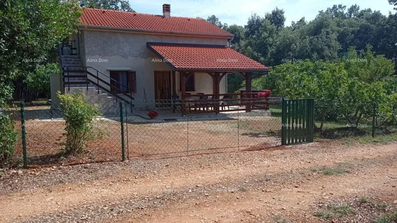 Kleines Haus mit rotem Ziegeldach, überdachter Terrasse und Außentreppe, umgeben von viel Grün in ländlicher Umgebung.