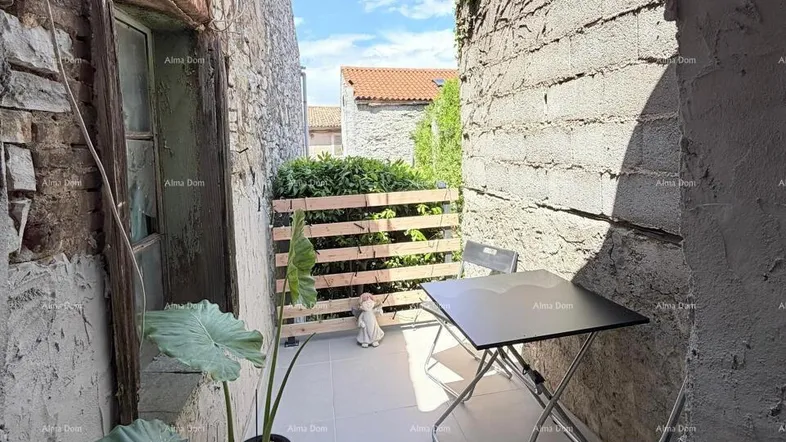 Schmale Außenterrasse zwischen Steinmauern mit kleinem Tisch, Stühlen und Topfpflanzen unter blauem Himmel.