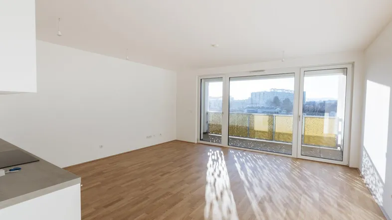 Heller offener Wohnbereich mit Parkettboden, großen Fenstern und Zugang zum Balkon.