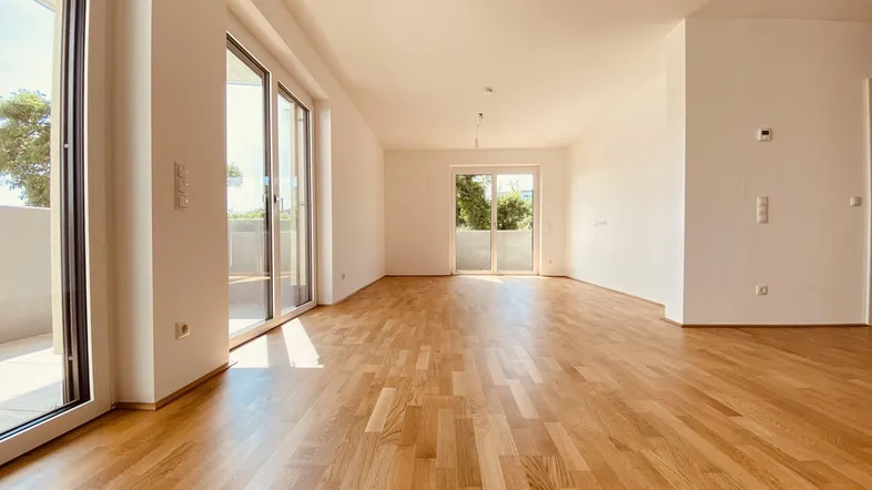 Helles, leeres Zimmer mit Parkettboden und großen Schiebetüren, die auf einen Balkon führen.