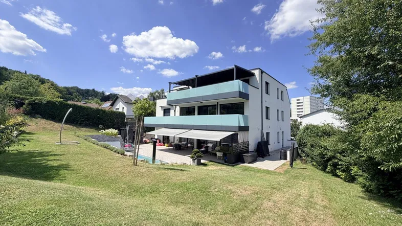 Modernes Wohnhaus mit mehreren Balkonen, großer Terrasse und weitläufigem Gartenbereich unter blauem Himmel.